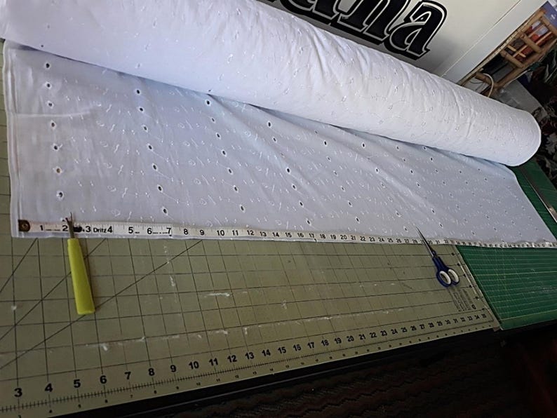 Pu&ograve; includere: Un rotolo di tessuto occhiellato bianco con un motivo ripetitivo di piccoli ritagli circolari. Un metro a nastro &egrave; posizionato sul tessuto, mostrando una larghezza di circa 71 cm. Sono presenti un taglia-asole e un paio di forbici blu.