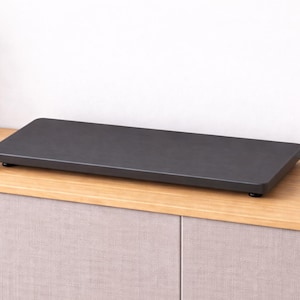 May include: A rectangular, dark gray serving tray with rounded corners sits on a light wood surface. The tray is elevated by small black feet. A small green plant in a gray pot is visible on the left.