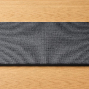 May include: A rectangular, dark gray mat with rounded corners. The mat has a textured surface and is placed on a light brown wooden surface. The mat appears to be made of a woven fabric.