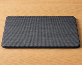 May include: A rectangular, dark gray mat with rounded corners. The mat has a textured surface and is placed on a light brown wooden surface. The mat appears to be made of a woven fabric.