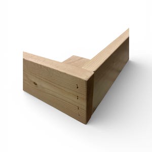 May include: A corner section of unfinished wooden frame, showcasing the natural grain and color of the wood. The frame is constructed with a mortise and tenon joint, and small nail holes are visible. The design suggests a furniture component or a structural element.