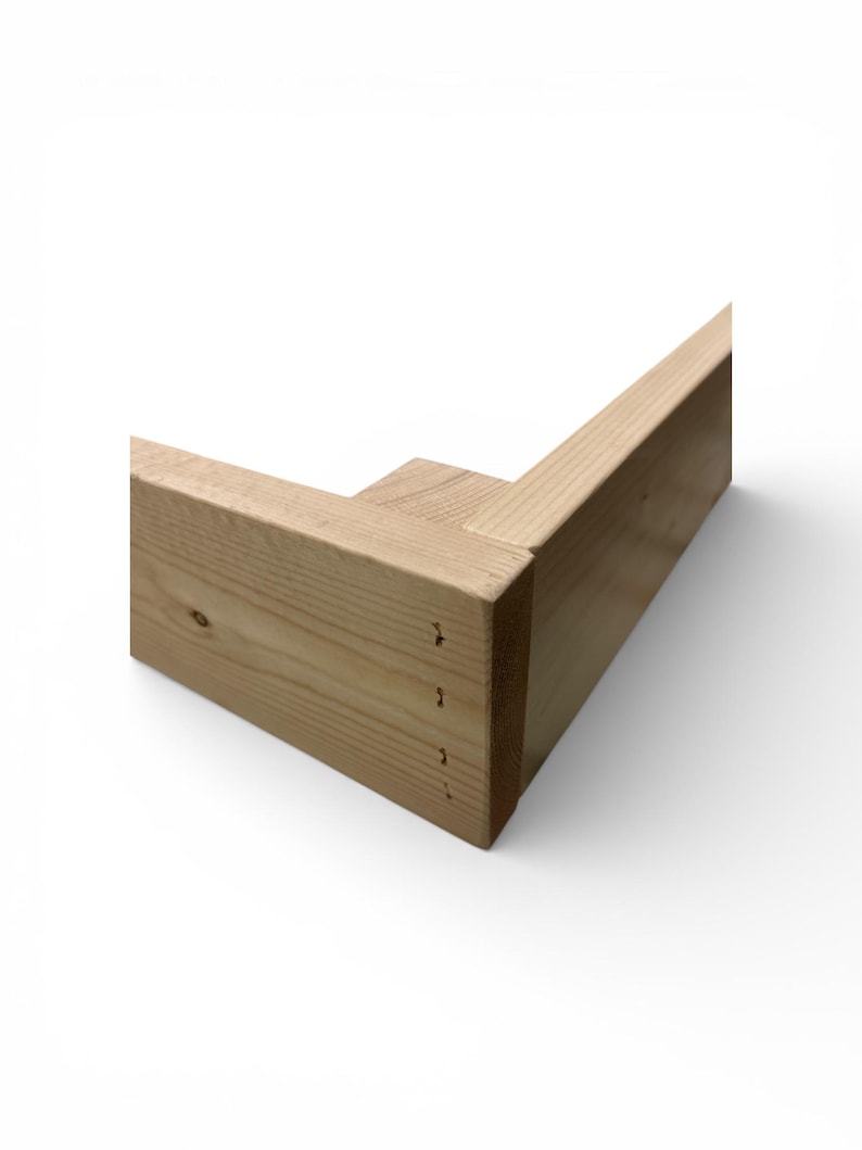May include: A corner section of unfinished wooden frame, showcasing the natural grain and color of the wood. The frame is constructed with a mortise and tenon joint, and small nail holes are visible. The design suggests a furniture component or a structural element.
