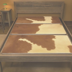 May include: A wooden bed frame with a headboard and three rectangular cowhide-patterned mattress pads. The pads feature a mix of brown and cream colors, creating a natural, rustic aesthetic. The bed is set against a neutral-toned wall.