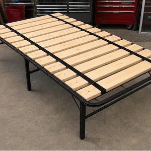 May include: A rectangular bed frame featuring a black metal base and a series of light-colored wooden slats arranged horizontally. The frame is supported by multiple black metal legs and includes two black straps running across the wooden slats.