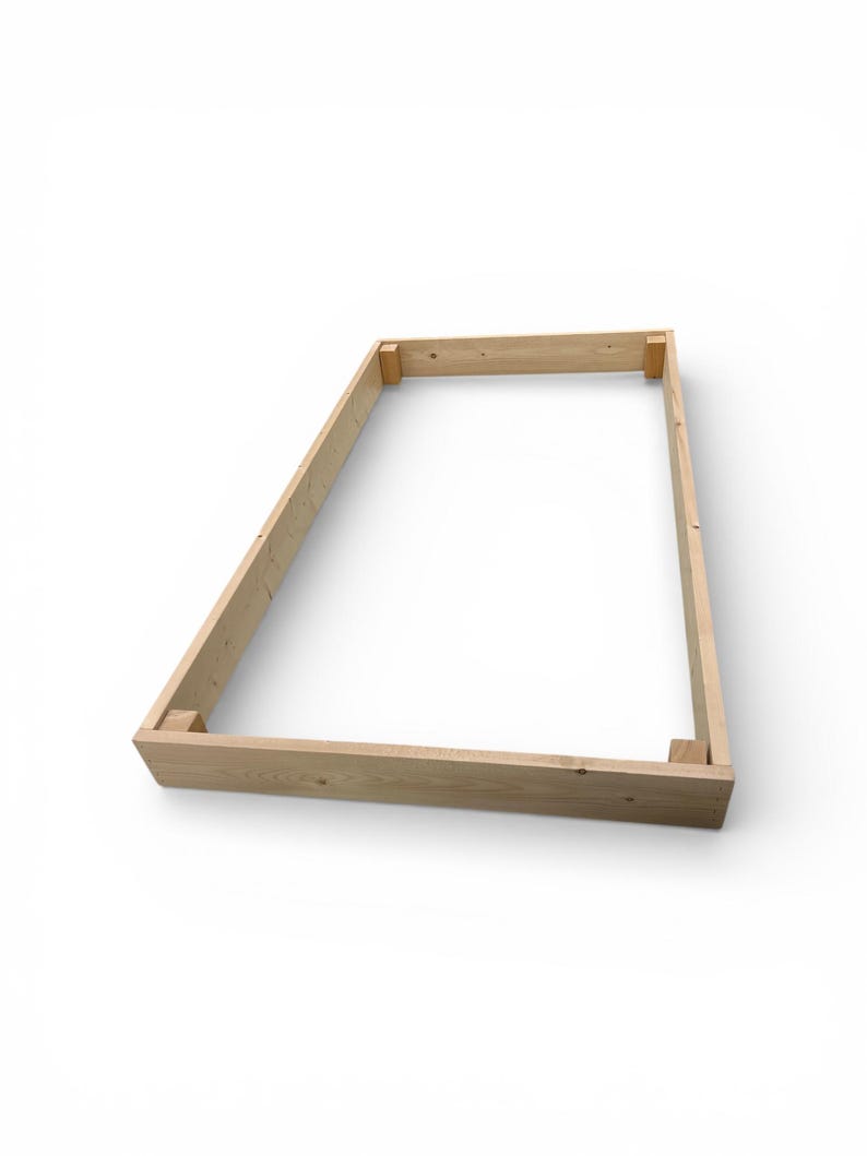 May include: A rectangular wooden frame, constructed from light-colored lumber, is presented against a white backdrop. The frame is assembled with corner joints, showcasing the natural wood grain and texture. The design suggests a potential for a raised garden bed or similar structure.