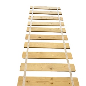 May include: A wooden slat bed frame with white straps. The slats are made of natural wood and are spaced evenly apart. The straps are attached to the slats and are used to secure the frame to the bed.