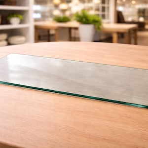 May include: A rectangular glass shelf with a polished edge, resting on a light brown wooden table. The glass reflects the surrounding environment, creating a transparent appearance. The shelf is designed for display or storage.
