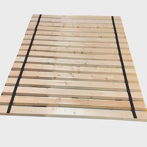 May include: A wooden bed frame with multiple horizontal wooden slats. The frame is made of light-colored wood and is held together by two black straps running vertically along the sides. The bed frame is designed to support a mattress.