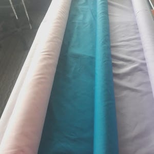 Pu&ograve; includere: Tre lunghi rotoli di tessuto disposti fianco a fianco. Il rotolo a sinistra &egrave; rosa pallido, il rotolo centrale &egrave; di un vibrante blu ottanio e il rotolo a destra &egrave; di un delicato color lavanda. Ogni rotolo &egrave; avvolto strettamente e si estende.