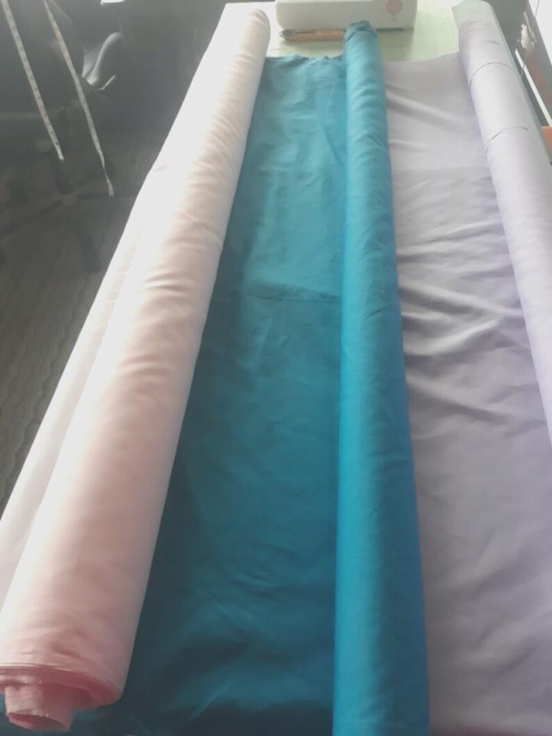 Pu&ograve; includere: Tre lunghi rotoli di tessuto disposti fianco a fianco. Il rotolo a sinistra &egrave; rosa pallido, il rotolo centrale &egrave; di un vibrante blu ottanio e il rotolo a destra &egrave; di un delicato color lavanda. Ogni rotolo &egrave; avvolto strettamente e si estende.