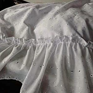 Pu&ograve; includere: Una balza per letto in tessuto bianco caratterizzata da un motivo in pizzo sangallo con piccoli fori circolari e dettagli ricamati. Il tessuto presenta un bordo arricciato che crea un aspetto stratificato e pieghettato lungo tutta la lunghezza.