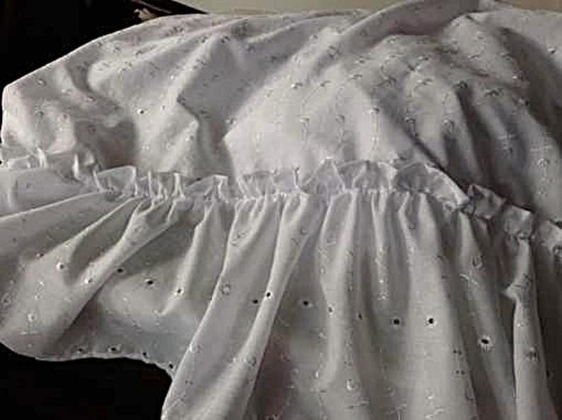 Pu&ograve; includere: Una balza per letto in tessuto bianco caratterizzata da un motivo in pizzo sangallo con piccoli fori circolari e dettagli ricamati. Il tessuto presenta un bordo arricciato che crea un aspetto stratificato e pieghettato lungo tutta la lunghezza.