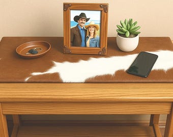 May include: A wooden framed photo of a couple wearing cowboy hats, a small succulent in a white pot, a brown leather tray with keys, and a black smartphone on a cowhide patterned table runner.