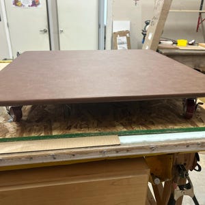 May include: A brown, rectangular table top with rounded corners, mounted on a wooden base with red caster wheels. The table is constructed from multiple layers of wood and other materials, suitable for various uses.