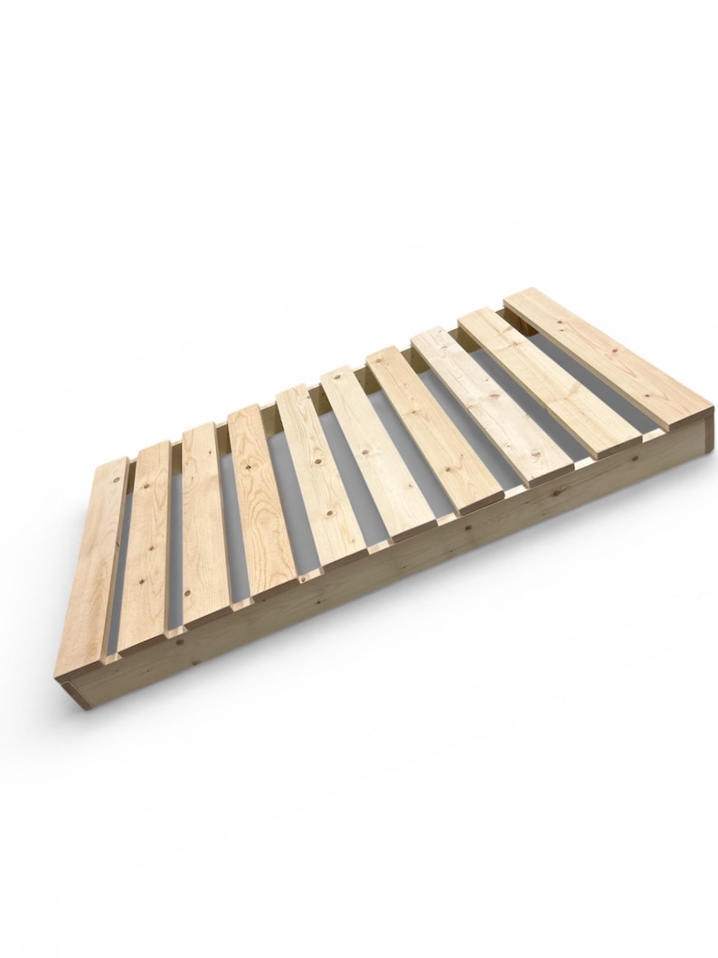 May include: Angled wooden bed frame with a slatted design. The frame is constructed from light-colored wood and features evenly spaced slats. The design is simple and modern, suitable for a minimalist bedroom aesthetic.