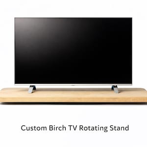 May include: A flat-screen TV with a black screen sits on a light-colored, rectangular birch wood stand. The stand has a rounded edge and is supported by silver legs. The text "Custom Birch TV Rotating Stand" is below the stand.