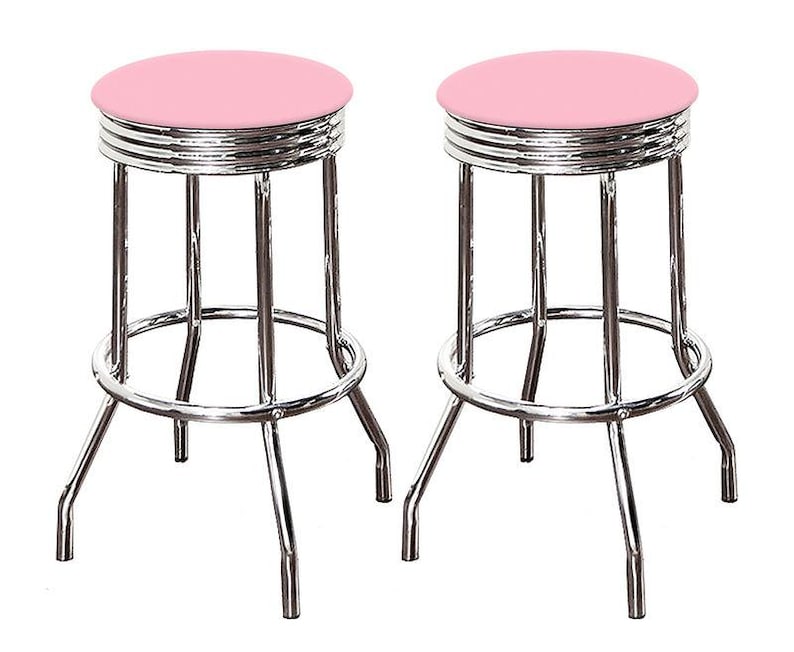 May include: Two retro-style bar stools. Each stool has a round, light pink seat and a chrome-finished metal frame. The frame features a footrest and four legs. The seats are approximately 12 inches in diameter.