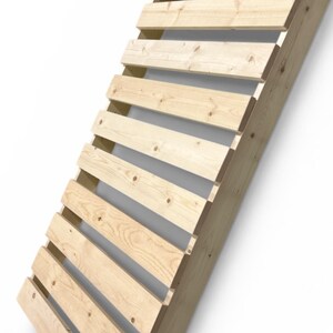 May include: Angled wooden structure with horizontal slats. The light-colored wood is constructed with visible joints and knots. The design appears to be a slatted backrest or decorative element, leaning against a white wall.