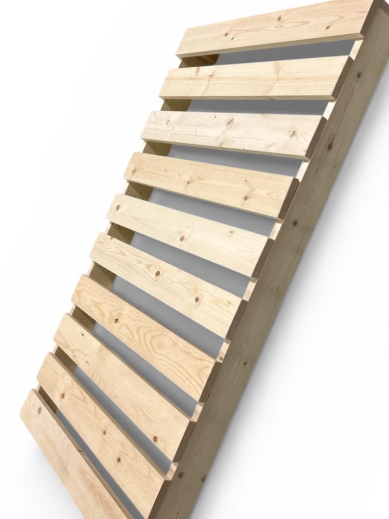 May include: Angled wooden structure with horizontal slats. The light-colored wood is constructed with visible joints and knots. The design appears to be a slatted backrest or decorative element, leaning against a white wall.
