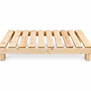 May include: A low-profile bed frame constructed from light-colored natural wood planks. The rectangular platform features a slatted surface design with evenly spaced horizontal boards supported by sturdy wooden legs at each of the four corners.