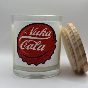 May include: A glass candle jar with a white label featuring a red and white Nuka Cola bottle cap design. The candle jar has a wooden lid.