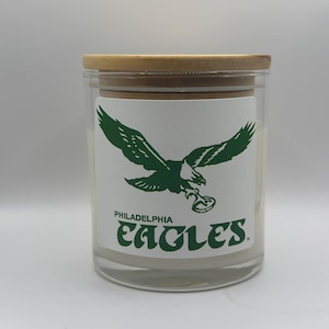May include: A clear glass candle jar with a wooden lid. The jar features a white label with a green Philadelphia Eagles logo and text. The candle is a home decor item.