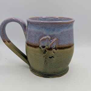 May include: A handmade ceramic mug with a sculpted floral design. The mug features a blue and green glaze, with a brown handle. The mug is approximately 4 inches tall and 3 inches in diameter, perfect for coffee or tea.