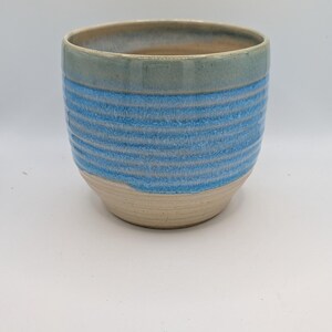 May include: A ceramic planter with a light blue and beige color scheme. The upper rim is a muted green-blue, transitioning to horizontal blue stripes. The base is a natural beige color. The planter has a rounded shape.