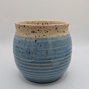 May include: A ceramic pot with a rounded shape and a wide opening. The pot features a light blue, textured exterior with horizontal lines. The rim is a speckled beige color. This decorative pot is suitable for plants or as a kitchen accent.