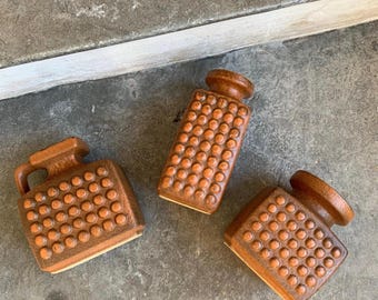Può includere: Tre contenitori in ceramica marrone con una superficie strutturata caratterizzata da punti circolari in rilievo. Un contenitore è rettangolare con manico, mentre gli altri due sono vasi quadrati più piccoli con aperture circolari piatte.