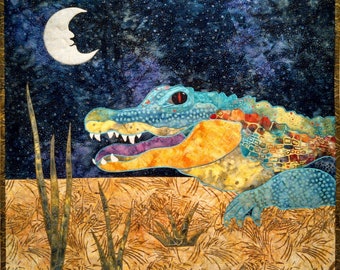 May include: A colorful fabric art piece depicting an alligator under a starry night sky with a crescent moon. The alligator is in shades of blue, orange, and yellow, with a red eye and open mouth. The background features tall grass.