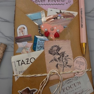 May include: A brown paper-wrapped parcel adorned with stickers, a pen with "UNFUCKHITHABLE" text, and a "Book Review" note. Embellishments include a rose illustration, a mushroom sticker, and a "FOCUS ON THE GOOD" tag. The package is tied with string.
