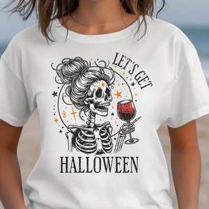Puede incluir: Camiseta blanca con una ilustración de esqueleto sosteniendo una copa de vino. El esqueleto tiene un peinado de moño y está rodeado de estrellas y el texto "LET'S GET HALLOWEEN". El diseño es en negro, naranja y blanco.