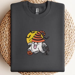 May include: Dark gray sweatshirt featuring a Halloween-themed embroidered design. The design includes a cartoon ghost wearing a witch hat, a full moon, bats, and a pumpkin. The sweatshirt is displayed on a woven, circular surface.