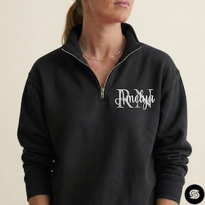 May include: Black quarter-zip sweatshirt with the name "Amelya" in a script font on the chest. The sweatshirt has a silver zipper and a high collar. The model is wearing a gold necklace.