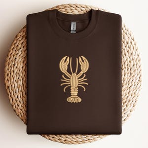 May include: A dark brown sweatshirt with a gold embroidered lobster design. The sweatshirt is folded and placed on a round woven mat. The lobster design is centered on the front of the shirt.