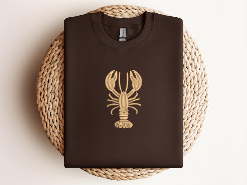 May include: A dark brown sweatshirt with a gold embroidered lobster design. The sweatshirt is folded and placed on a round woven mat. The lobster design is centered on the front of the shirt.