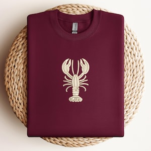 May include: Burgundy-colored sweatshirt with a cream-colored embroidered lobster design. The sweatshirt is folded and placed on a round woven mat. The lobster design is centered on the front of the shirt.