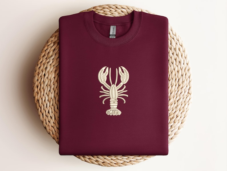 May include: Burgundy-colored sweatshirt with a cream-colored embroidered lobster design. The sweatshirt is folded and placed on a round woven mat. The lobster design is centered on the front of the shirt.