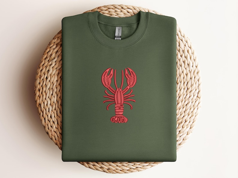 May include: An olive green sweatshirt with a red embroidered lobster design. The lobster is centered on the front of the shirt. The sweatshirt is folded and placed on a woven, circular mat.