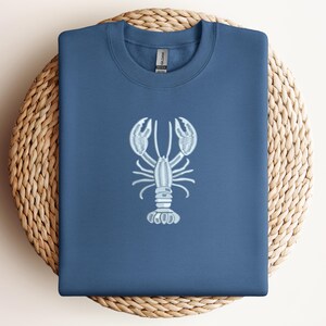 May include: A navy blue sweatshirt with a light blue embroidered lobster design. The sweatshirt is folded and placed on a round, woven wicker placemat. The lobster design is centered on the front of the shirt.