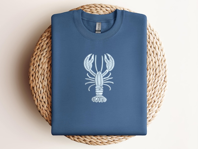 May include: A navy blue sweatshirt with a light blue embroidered lobster design. The sweatshirt is folded and placed on a round, woven wicker placemat. The lobster design is centered on the front of the shirt.