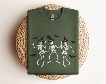 May include: A green sweatshirt with a graphic of three skeletons wearing witch hats and holding brooms. The skeletons are dancing and there are bats flying around them.
