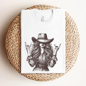 May include: White t-shirt featuring a black and white illustration of a woman wearing a cowboy hat and making a rock and roll hand gesture. The design includes long flowing hair and a belt. The shirt is folded on a woven, circular surface.