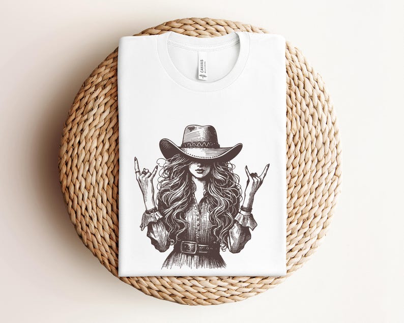 May include: White t-shirt featuring a black and white illustration of a woman wearing a cowboy hat and making a rock and roll hand gesture. The design includes long flowing hair and a belt. The shirt is folded on a woven, circular surface.
