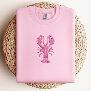 May include: A light pink sweatshirt with a pink embroidered lobster design. The lobster is centered on the front of the shirt. The sweatshirt is displayed on a round, woven, light brown surface.