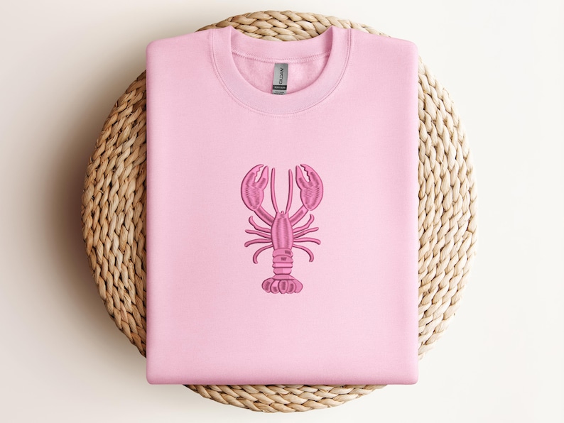 May include: A light pink sweatshirt with a pink embroidered lobster design. The lobster is centered on the front of the shirt. The sweatshirt is displayed on a round, woven, light brown surface.
