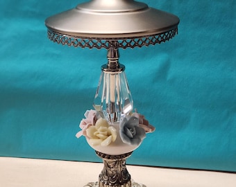 May include: A silver and glass decorative table lamp with a crystal finial and a floral design. The lamp has a round base and a wide, flat top.