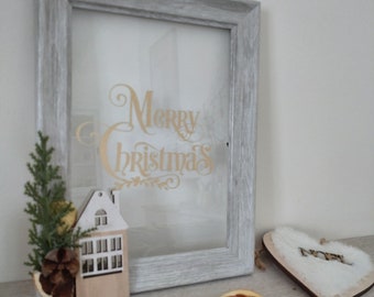 May include: A gray wooden frame with a glass front, featuring the words "Merry Christmas" in gold lettering. A small wooden house with windows and a pine sprig with a pine cone are in the frame. The frame is on a wooden surface with dried orange slices and star anise.