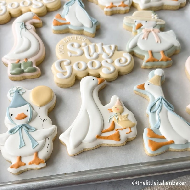 May include: Assortment of decorated sugar cookies featuring goose designs. Cookies include geese with hats, bows, and boots, and the phrase "Our Little Silly Goose". The cookies are decorated with white, blue, orange, and yellow icing.
